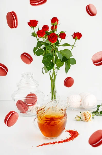 Flying food and still life concept. Breakfast desserts on white background with macaroons, bouquet of red roses and splashing tea.