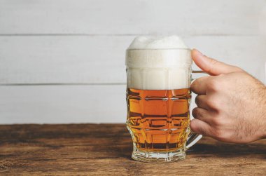 Ahşap masada köpüklü bira bardağını tutan bir erkek el. Tonu ayarlanmış resim, alanı kopyala