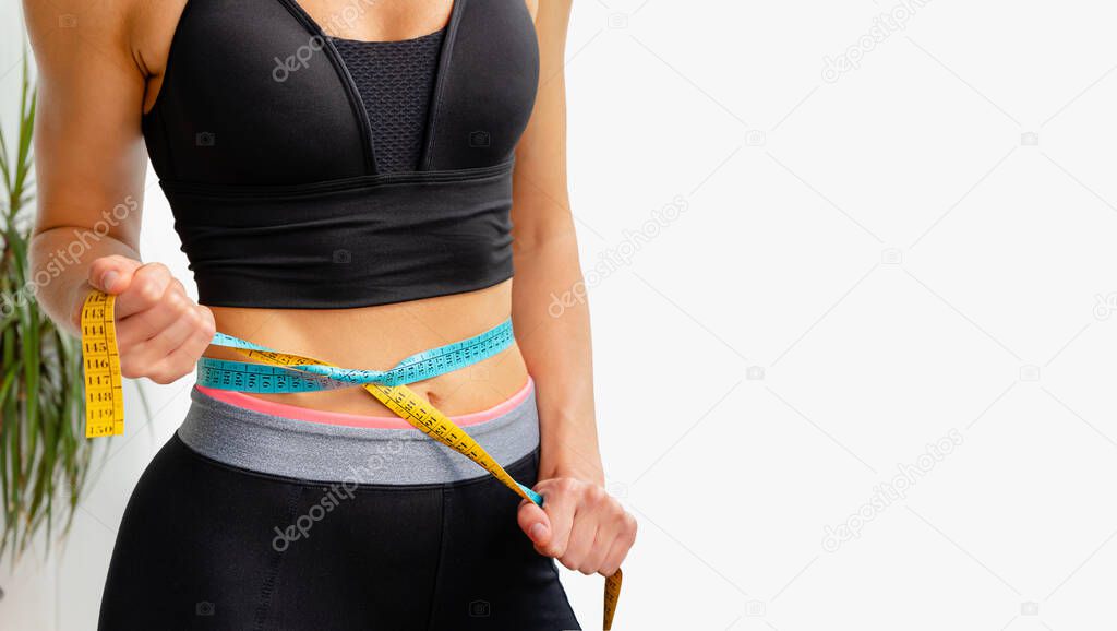 Young slim woman tightens measuring tape on her waist. Sporty lifestyle and weight loss concept.