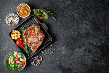 Izgara domuz bifteği, dökme demir tavada, baharatlar ve sebzeler koyu arka planda. Boşluğu kopyala, üst görünüm