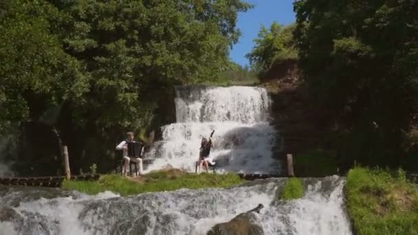 Twee muzikanten Pianist en bandurist spelen op de rotsen bij de waterval — Stockvideo