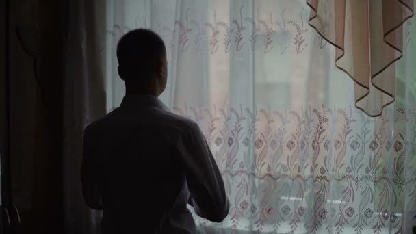 Молодий чоловік захоплюється видом з вікна вдома і одягає сорочку. Повільний рух — стокове відео