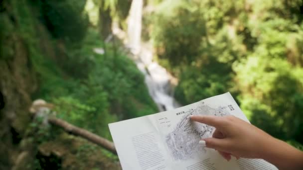 Turista femenina con una mochila revisando mapa de papel cerca de la cascada de montaña — Vídeos de Stock