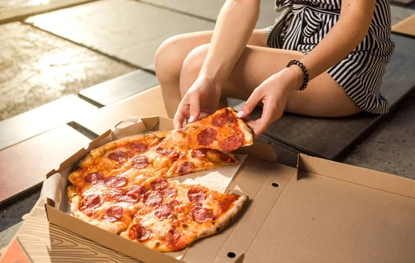 Happy young woman holding hot pizza in box, seat outdoor in park