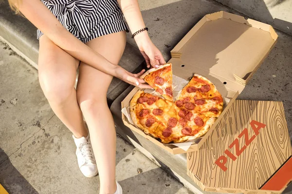 Happy young woman holding hot pizza in box, seat outdoor in park