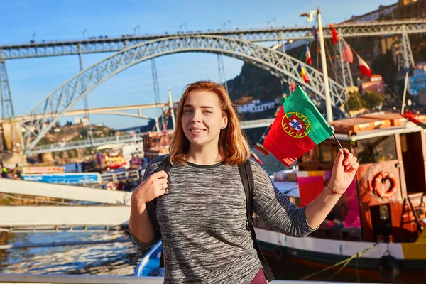 Mujer joven viajera de pie con bandera portuguesa, disfrutando de hermosas vistas del paisaje urbano en el río Duero, puente y barcos durante la luz de la mañana en Oporto, Portugal — Foto de Stock