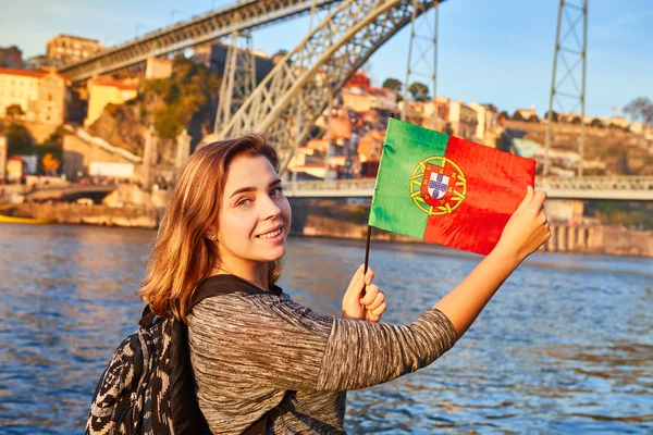Ung kvinna turist med portugisiska flaggan njuter vackra landskap syn på den gamla staden med floden och berömda järn bron Dom Luiz under solnedgången i staden Porto, Portugal — Stockfoto
