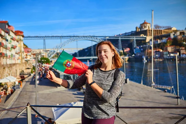 Mujer joven viajera de pie con bandera portuguesa, disfrutando de hermosas vistas del paisaje urbano en el río Duero, puente y barcos durante la luz de la mañana en Oporto, Portugal —  Fotos de Stock