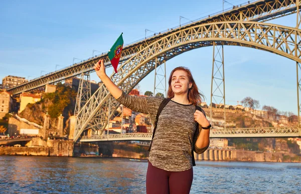 Joven turista con bandera portuguesa disfrutando de una hermosa vista del paisaje en el casco antiguo con río y famoso puente de hierro Dom Luiz durante la puesta de sol en la ciudad de Oporto, Portugal — Foto de Stock