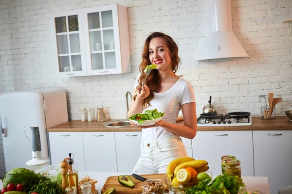 실내에서 녹색 신선한 재료와 아름다운 부엌에서 샐러드를 먹는 젊은 행복한 여성. 건강한 음식 개념 — 스톡 사진