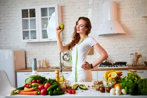 Joven mujer feliz comiendo manzana en la hermosa cocina con ingredientes frescos verdes en el interior. Comida saludable y concepto de dieta. Perder peso —  Fotos de Stock