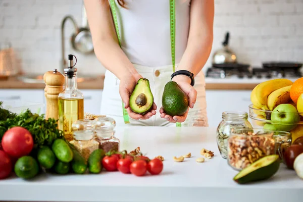 Νεαρή ευτυχισμένη γυναίκα κρατώντας το αβοκάντο για να κάνετε σαλάτα στην όμορφη κουζίνα με πράσινα φρέσκα υλικά σε εσωτερικούς χώρους. Υγιεινά φαγητά και δίαιτες. Χάνοντας βάρος — Φωτογραφία Αρχείου
