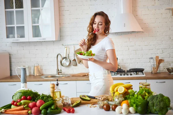 Mladá šťastná žena jedla salát v nádherné kuchyni se zelenými čerstvými ingrediencemi uvnitř. Koncepce zdravého jídla — Stock fotografie