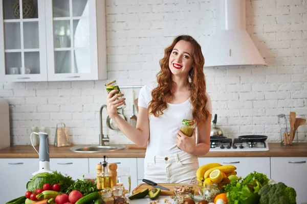 Jovem mulher bebendo smoothie verde e água doce com pepino, limão, folhas de hortelã na mesa da cozinha com frutas e legumes. Conceito de alimentação saudável. Vegan refeição e menu de desintoxicação — Fotografia de Stock