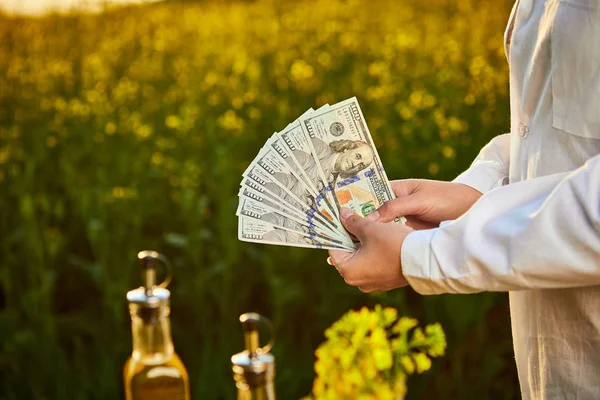 Agronómus nő vagy gazdálkodó gazdaság pénz dollár bankjegy virágzó termesztett repce mező-és repcemag-olaj a háttérben — Stock Fotó
