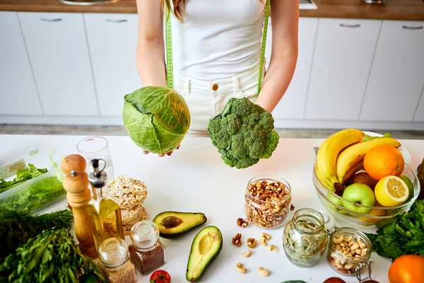 Νέα ευτυχισμένη γυναίκα κρατώντας μπρόκολο και λάχανο στην όμορφη κουζίνα με πράσινα φρέσκα υλικά σε εσωτερικούς χώρους. Υγιεινά φαγητά και δίαιτες. Χάνοντας βάρος — Φωτογραφία Αρχείου