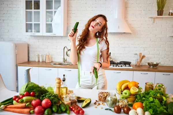 Mladá šťastná žena jedla okurku za výrobu salátu v nádherné kuchyni se zelenými čerstvými ingrediencemi uvnitř. Zdravá strava a dieting koncepce. Uvolňující váha — Stock fotografie