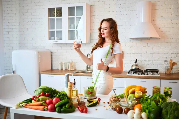 Mladá žena pije čerstvou vodu ze skla a v kuchyni ukazuje palce. Zdravý životní styl a jídlo. Zdraví, krása, dietní koncepce. — Stock fotografie