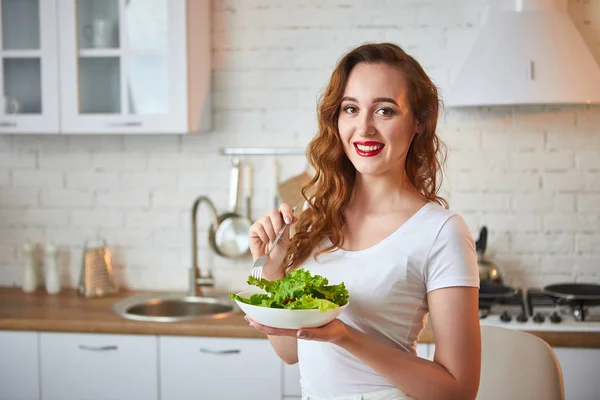 Fiatal boldog nő étkezési saláta a gyönyörű konyhában, zöld friss alapanyagokból beltérben. Egészséges táplálkozás koncepció — Stock Fotó
