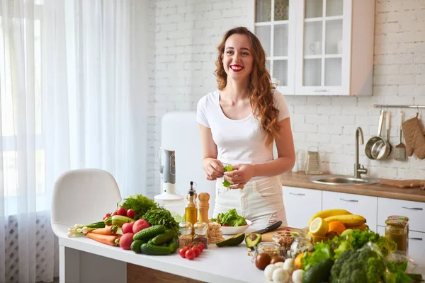 Mladá šťastná žena připravuje chutné saláty v nádherné kuchyni se zelenými čerstvými ingrediencemi uvnitř. Zdravá strava a dieting koncepce. Uvolňující váha — Stock fotografie