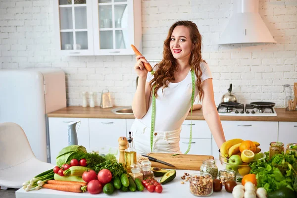 Mladá šťastná žena držící v krásné kuchyni mrkev s čerstvými zelenými ingrediencemi uvnitř. Zdravá strava a dieting koncepce. Uvolňující váha — Stock fotografie