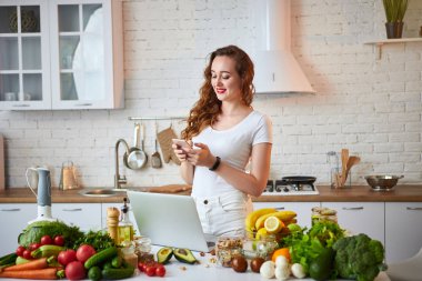 Genç güzel kadın akıllı telefon konuşuyor ve modern mutfakta yemek yaparken bir dizüstü bilgisayar kullanarak. Sağlıklı gıda ve Diyet kavramı. Kilo Verme