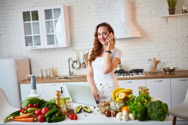 Genç güzel kadın akıllı telefon konuşuyor ve modern mutfakta yemek yaparken bir dizüstü bilgisayar kullanarak. Sağlıklı gıda ve Diyet kavramı. Kilo Verme