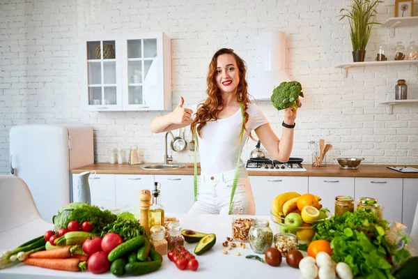 브로콜리를 들고 실내에서 녹색 신선한 재료와 아름다운 부엌에서 엄지 손가락을 보여주는 젊은 행복한 여성. 건강한 음식과 다이어트 개념. 체중 감량 — 스톡 사진