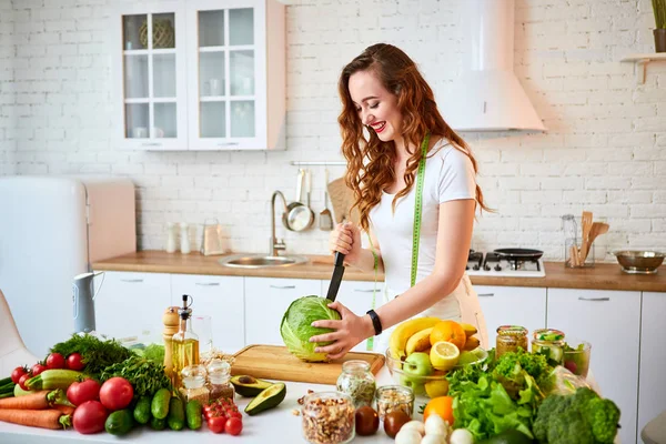 Mladá šťastná žena krájí zelí na výrobu salátu v nádherné kuchyni se zelenými čerstvými ingrediencemi uvnitř. Zdravá strava a dieting koncepce. Uvolňující váha — Stock fotografie