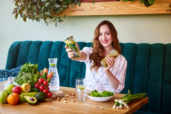 Young woman drinking smoothie and fresh water in the beautiful interior with green flowers on the background and fresh fruits and vegetables on the table. Healthy eating concept. Vegan meal and detox menu