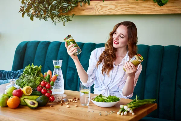 Young woman drinking smoothie and fresh water in the beautiful interior with green flowers on the background and fresh fruits and vegetables on the table. Healthy eating concept. Vegan meal and detox menu