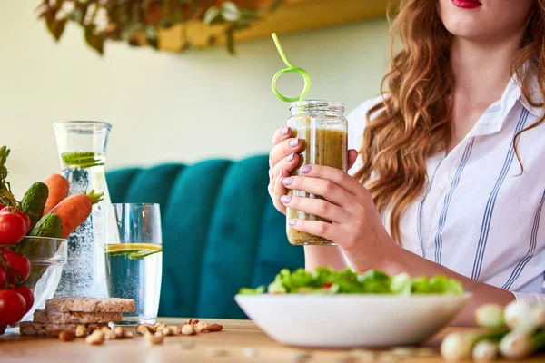 Νεαρή γυναίκα που πίνει χυμό στο όμορφο εσωτερικό με πράσινα λουλούδια στο φόντο και φρέσκα φρούτα και λαχανικά στο τραπέζι. Έννοια της υγιεινής διατροφής. Χορτοφαγικό γεύμα και μενού αποτοξίνωσης — Φωτογραφία Αρχείου