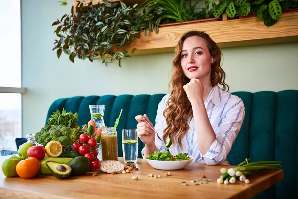 배경에 녹색 꽃과 테이블에 신선한 재료와 아름다운 인테리어에 샐러드를 먹고 젊은 행복한 여성. 건강한 음식 개념 — 스톡 사진