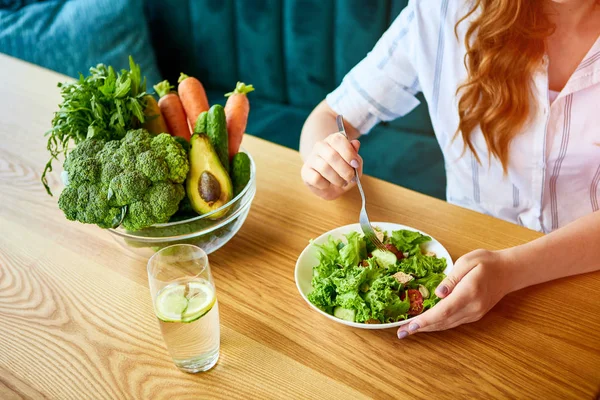 Νέα ευτυχισμένη γυναίκα τρώγοντας σαλάτα στο όμορφο εσωτερικό με πράσινα λουλούδια στο φόντο και φρέσκα υλικά στο τραπέζι. Υγιεινή διατροφή — Φωτογραφία Αρχείου