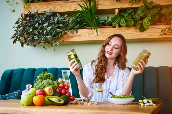 Young woman drinking smoothie and fresh water in the beautiful interior with green flowers on the background and fresh fruits and vegetables. Healthy eating concept. Vegan meal and detox menu
