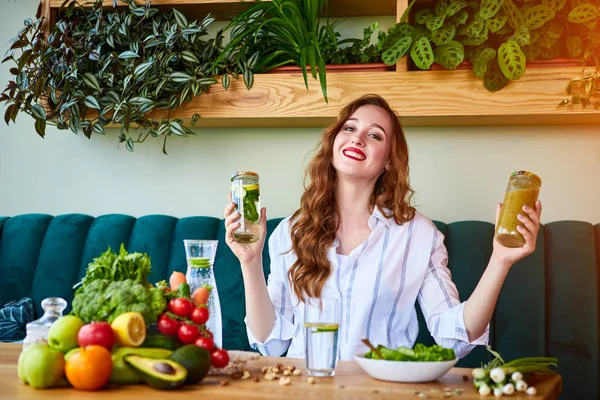 Young woman drinking smoothie and fresh water in the beautiful interior with green flowers on the background and fresh fruits and vegetables. Healthy eating concept. Vegan meal and detox menu