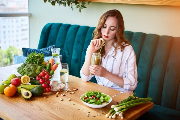 Νέα γυναίκα πίνοντας χυμό στο όμορφο εσωτερικό με πράσινα λουλούδια στο φόντο και φρέσκα φρούτα και λαχανικά. Έννοια της υγιεινής διατροφής. Χορτοφαγικό γεύμα και μενού αποτοξίνωσης — Φωτογραφία Αρχείου