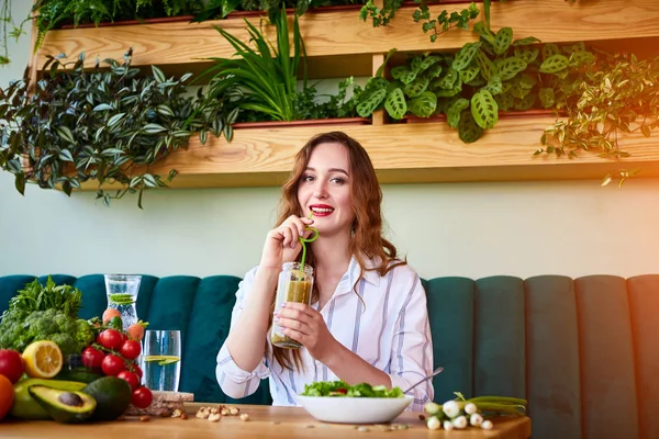 Young woman drinking smoothie in the beautiful interior with green flowers on the background and fresh fruits and vegetables. Healthy eating concept. Vegan meal and detox menu