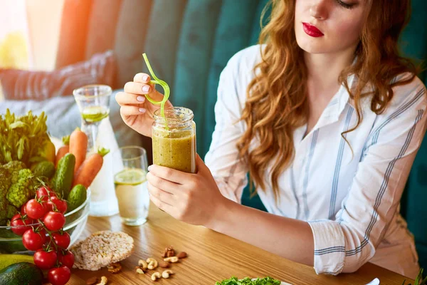 Young woman drinking smoothie in the beautiful interior with green flowers on the background and fresh fruits and vegetables. Healthy eating concept. Vegan meal and detox menu