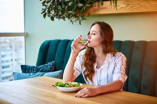 Young happy woman drinks water in the beautiful interior with green flowers on the background and fresh ingredients on the table. Healthy food concept