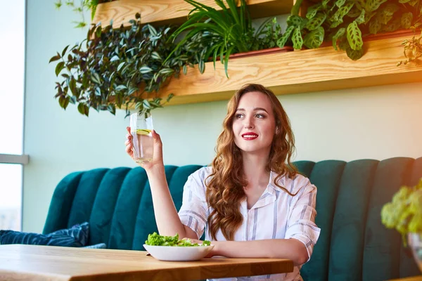 Young happy woman drinks water in the beautiful interior with green flowers on the background and fresh ingredients on the table. Healthy food concept