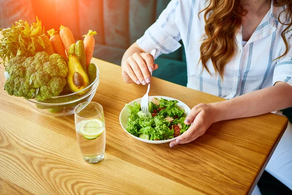 Νέα ευτυχισμένη γυναίκα τρώγοντας σαλάτα στο όμορφο εσωτερικό με πράσινα λουλούδια στο φόντο και φρέσκα υλικά στο τραπέζι. Υγιεινή διατροφή — Φωτογραφία Αρχείου