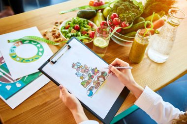 Tıbbi üniformalı kadın diyetisyen, arka planda yeşil ofiste farklı sağlıklı gıda malzemelerle oturan bir diyet planı üzerinde çalışan bant ölçümü ile. Kilo kaybı ve sağ beslenme konsepti