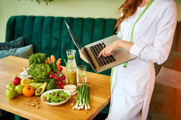 Woman dietitian in medical uniform with tape measure working with laptop on a diet plan standing near different healthy food ingredients in the green office on background. Weight loss and right nutrition concept