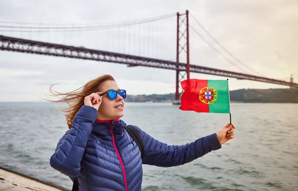 Młoda kobieta turysta trzymając flagę Portugalii w rękach i korzystających widok krajobrazu na słynnym Żelaznym mostem 25 kwietnia stoi z powrotem na nabrzeżu w Lizbonie miasta — Zdjęcie stockowe