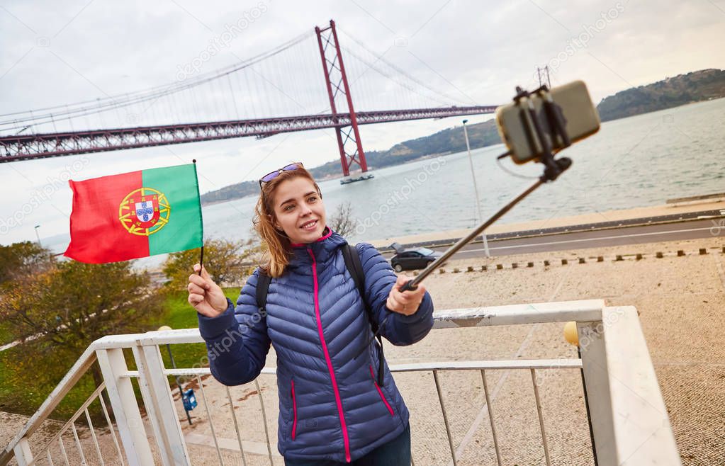 Young woman tourist take selfies on the background of famous iron 25th of April bridge and holding the flag of Portugal in hands  in Lisbon city