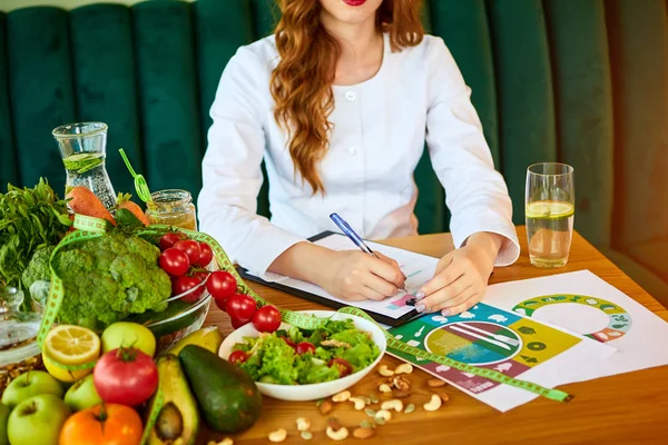 医療服を着た女性栄養士は、背景に緑のオフィスで異なる健康的な食材を持つダイエット計画に取り組んでテープメジャーを持っています。減量と右栄養の概念 — ストック写真