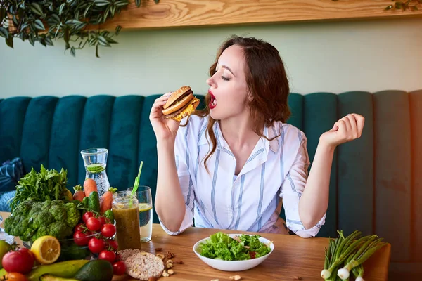 Beautiful young woman eating hamburger instead of salad in kitchen. Cheap junk food vs healthy diet — Stock Photo, Image