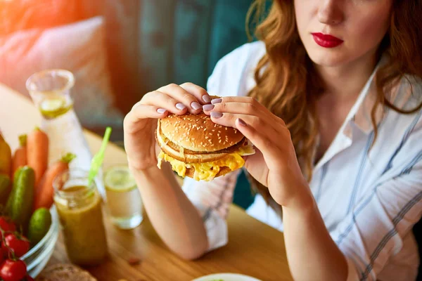 キッチンでサラダの代わりにハンバーガーを食べる美しい若い女性。安いジャンクフード対健康的な食事 — ストック写真