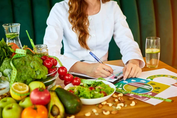 Woman dietitian in medical uniform with tape measure working on a diet plan sitting with different healthy food ingredients in the green office on background. Weight loss and right nutrition concept — Stock Photo, Image